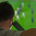 A man sitting on a couch watching a soccer game on his TV. He is holding the TV remote close to his head.