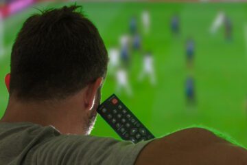 A man sitting on a couch watching a soccer game on his TV. He is holding the TV remote close to his head.