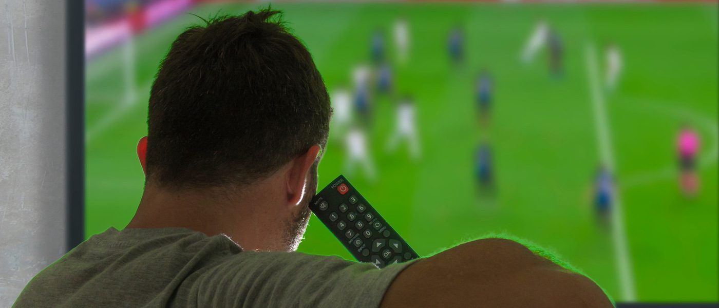 A man sitting on a couch watching a soccer game on his TV. He is holding the TV remote close to his head.
