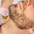 A bearded man in a robe leaning his head back as a technician uses a laser hair removal device against his throat.
