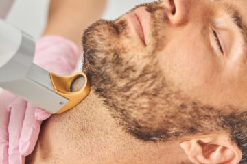 A bearded man in a robe leaning his head back as a technician uses a laser hair removal device against his throat.