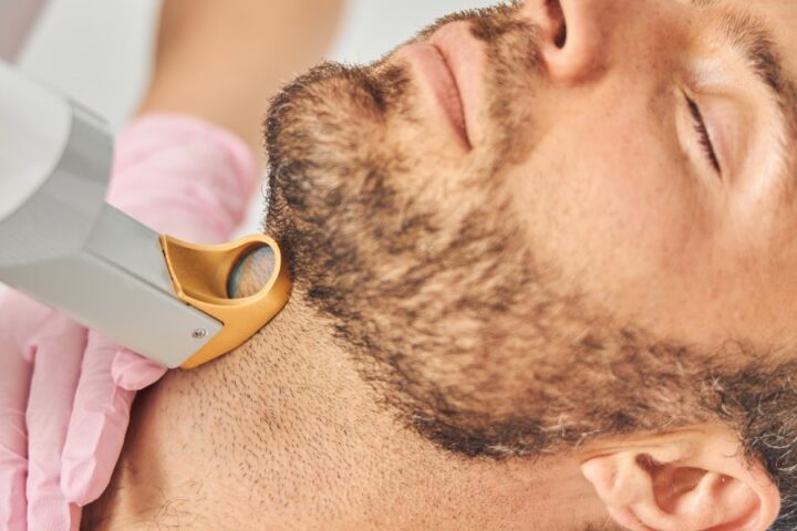 A bearded man in a robe leaning his head back as a technician uses a laser hair removal device against his throat.