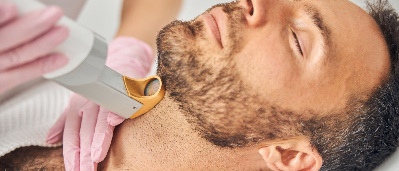A bearded man in a robe leaning his head back as a technician uses a laser hair removal device against his throat.