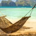 A brown woven hammock hanging between two tree in the shade. The hammock overlooks the beach of a lovely tropical island.