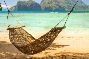 A brown woven hammock hanging between two tree in the shade. The hammock overlooks the beach of a lovely tropical island.