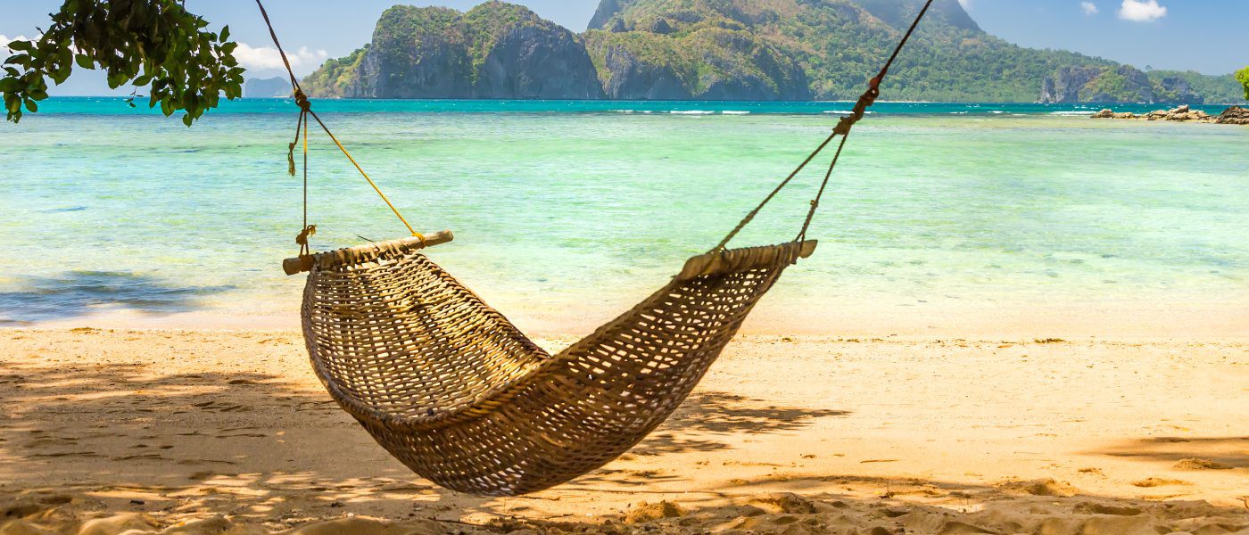 A brown woven hammock hanging between two tree in the shade. The hammock overlooks the beach of a lovely tropical island.