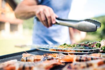 A man in a blue T-shirt, standing at his barbecue grill in his backyard, far away from the house. He grills various seafood.