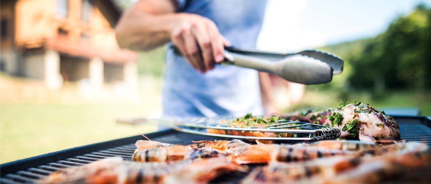 A man in a blue T-shirt, standing at his barbecue grill in his backyard, far away from the house. He grills various seafood.