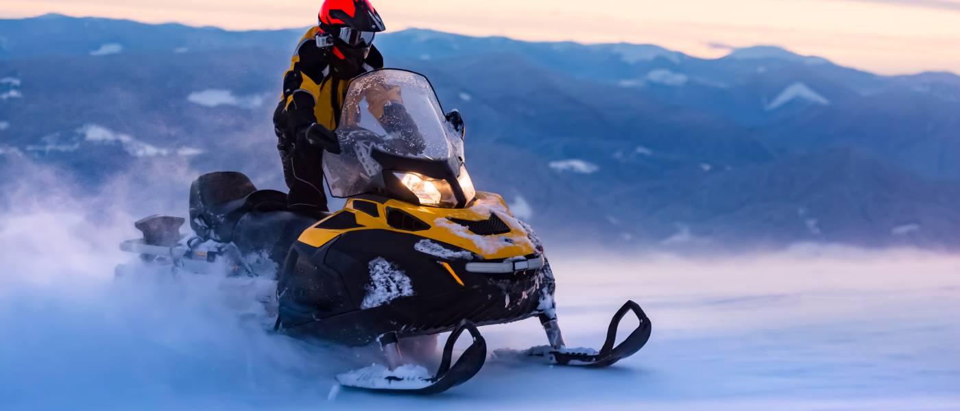 A man riding a black-and-yellow snowmobile on a mountain. Behind him are a mountain range and pastel sky at dusk.