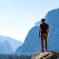 A man in khakis, a T-shirt, and a baseball cap stands on a rock overlooking midday scenery in Yosemite National Park.