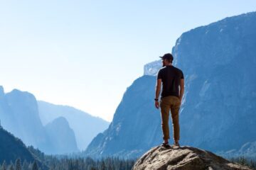 A man in khakis, a T-shirt, and a baseball cap stands on a rock overlooking midday scenery in Yosemite National Park.