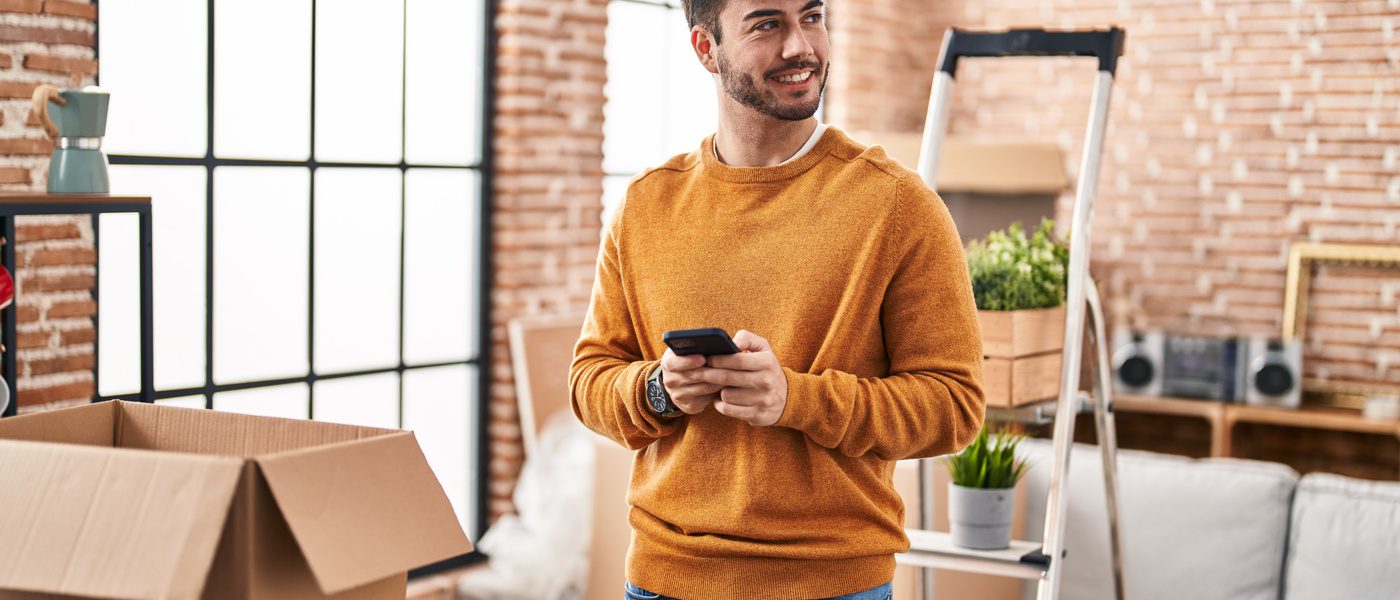 A young man wearing an orange sweater, smiling and standing in his new bachelor pad, surrounded by moving boxes.