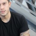 A young man with a lip piercing sitting on public steps. He is wearing a black T-shirt and false tattoo sleeves.