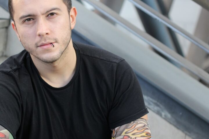 A young man with a lip piercing sitting on public steps. He is wearing a black T-shirt and false tattoo sleeves.