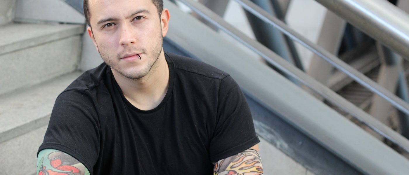 A young man with a lip piercing sitting on public steps. He is wearing a black T-shirt and false tattoo sleeves.