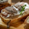 Thick slices of bruschetta bread sitting on a wooden cutting board. Each slice has lard topped with shaved black truffle.