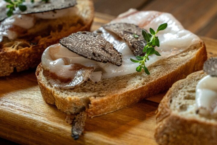 Thick slices of bruschetta bread sitting on a wooden cutting board. Each slice has lard topped with shaved black truffle.