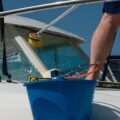 Someone standing on top of their boat using a brush to scrub the windshield. A blue bucket with more supplies is also on the deck.