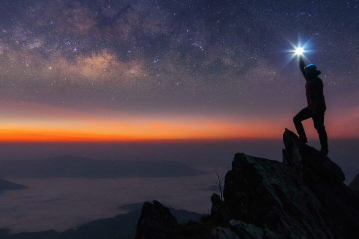 The silhouette of a man shining a flashlight while standing on a mountain's summit at dusk. The Milky Way is visible.