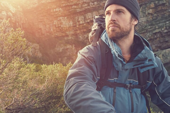 A man wearing a blue beanie and windbreaker jacket is outside hiking in the mountains and holding a paper map.