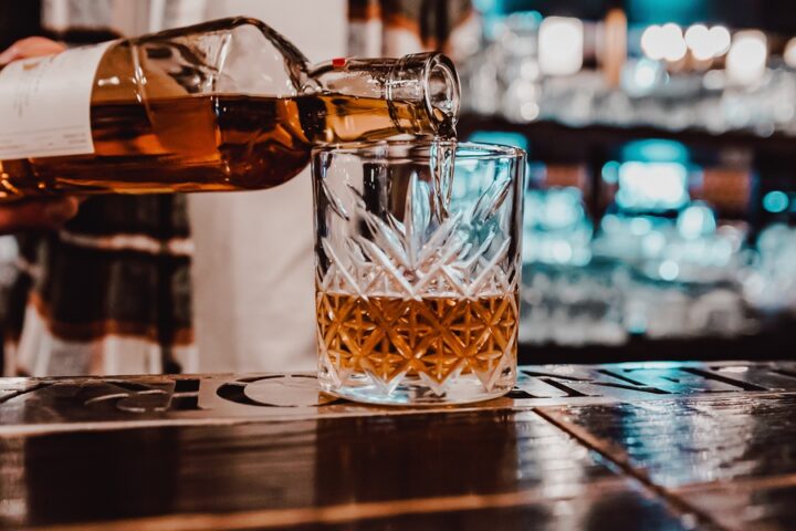 A bartender pours whiskey from a bottle into a rocks glass that sits on a wide bar top. Shelves of liquor sit behind him.