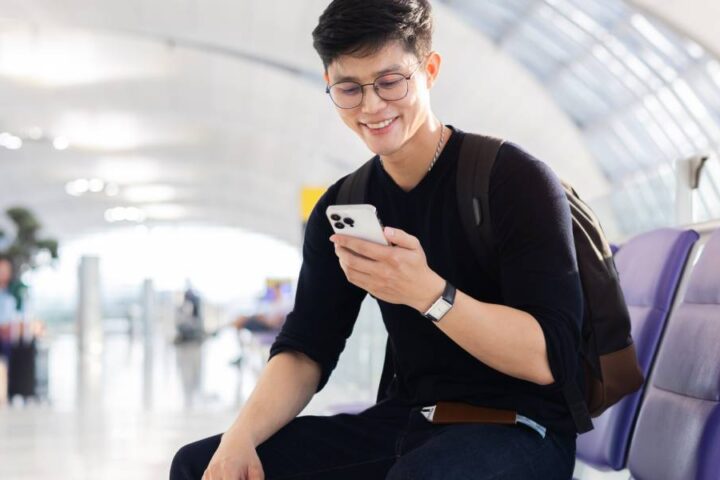 A young Asian man smiles and uses a smartphone while sitting in the airport near his departure area.