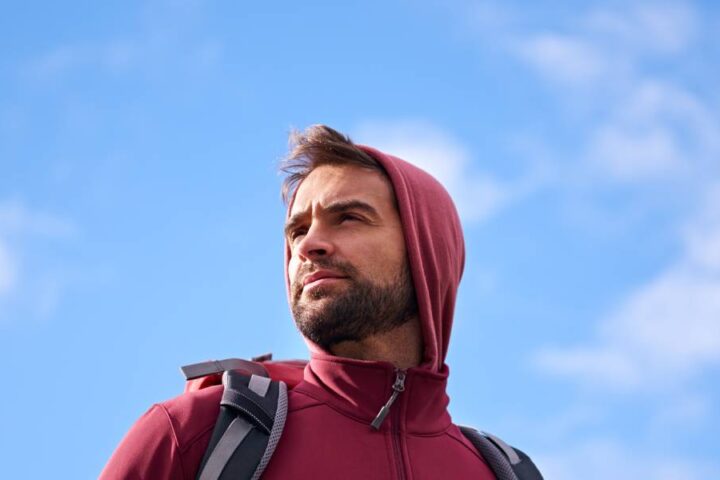 A man with a short beard and brown hair standing against a blue sky with white clouds while wearing outdoor gear.