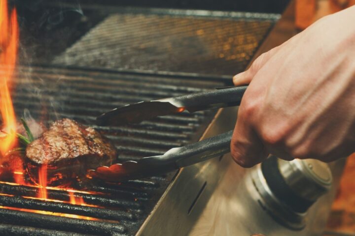 A man using metal tongs to grab a steak off a grill. The fire from under the grates is surrounding the charred meat.