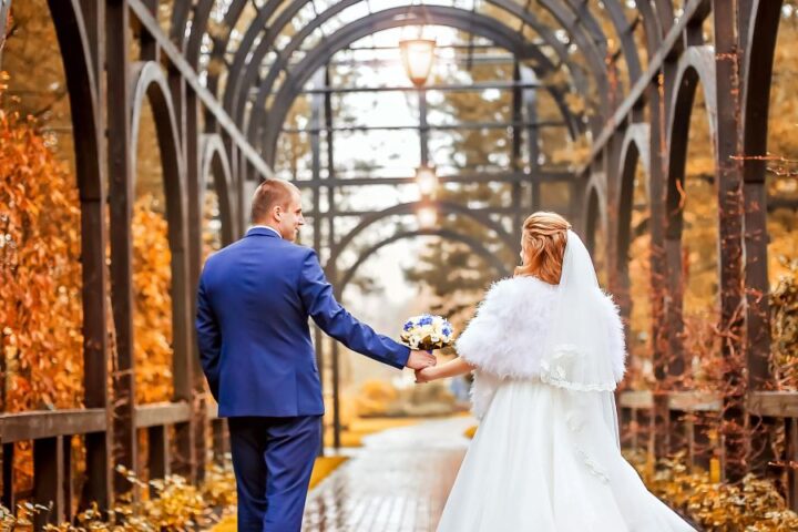 A groom and bride walk through an archway with orange fall leaves surrounding them. The groom reaches out for the bride.