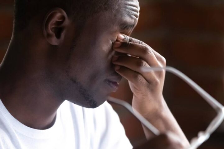 An exhausted man holding his white-framed glasses to the side so that he can rub his eyes with his free hand.
