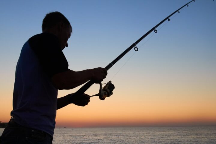 A fisherman casting a line into a lake as the sun sets in the background. He is holding the fishing pole with two hands.