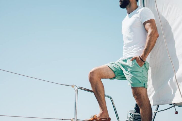 A man in turquoise shorts is standing on the edge of his boat and is enjoying the view of the ocean.