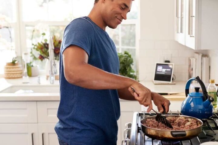 A young adult Black man is cooking on the stove, using a spatula and a frying pan. He's smiling while he cooks.