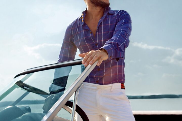 A man standing on the deck of a yacht. He's wearing a blue plaid button-up shirt and a pair of white slacks.