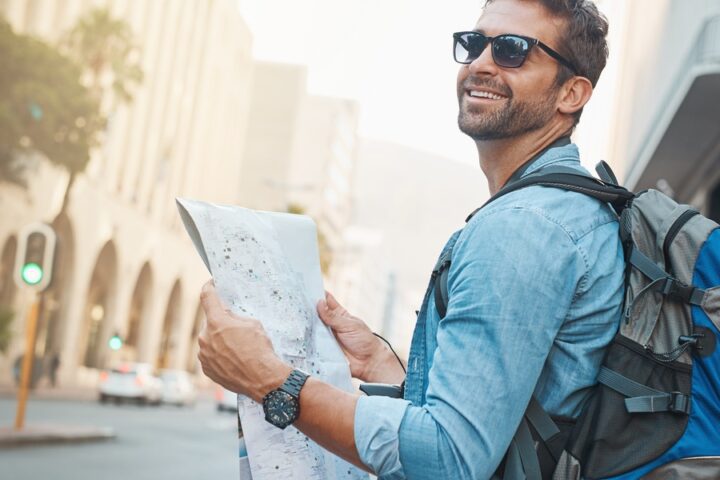 A traveler in his 30s smiles and looks up from a map as he navigates a city at day. He wears a backpack, watch, and sunglasses.