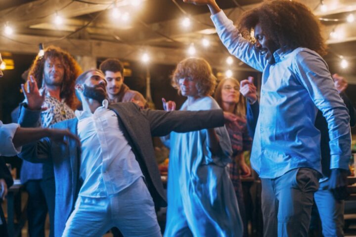 A bunch of happy wedding guests dancing under a large pergola decorated with string lights.