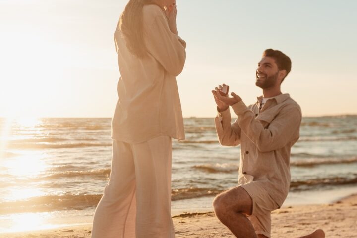 A man kneeling to propose to his surprised, happy girlfriend on the shoreline of a beach at sunset.