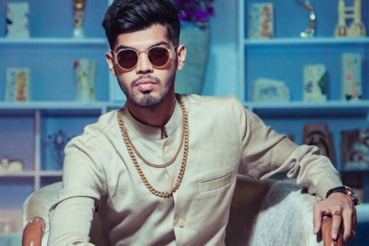 A man adorned with gold jewelry, including a necklace, bracelet, rings, and a watch, sitting on a chair in a living room.