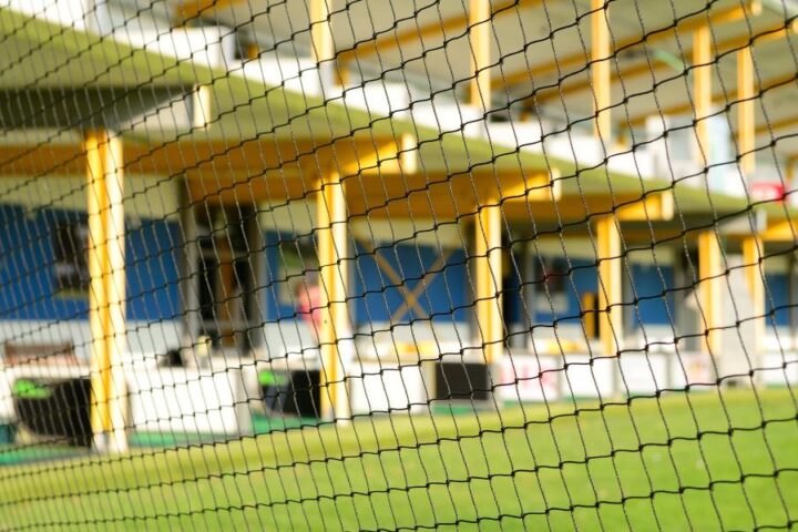 A close-up view of a net with a driving range visible through it. A man is taking a swing with a golf club.