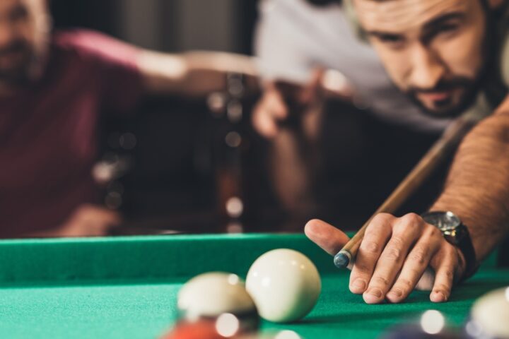 A man leans over a pool table with green felt, preparing to take his shot, as two male friends look on in the background.