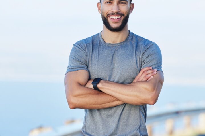 A young, healthy male athlete stands with crossed arms, smiling to the side of the road after a run.