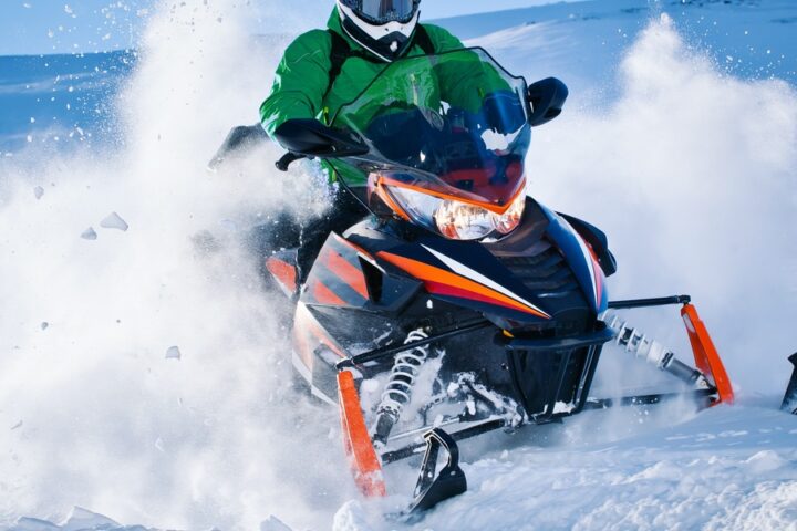 A person in a bright green jacket rides a black snowmobile, kicking up snow in a scenic, snowy mountain landscape.
