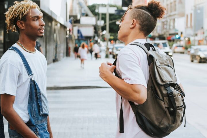 Two young men chat on a bustling city street; one wears overalls, the other a backpack, with urban life in the background.
