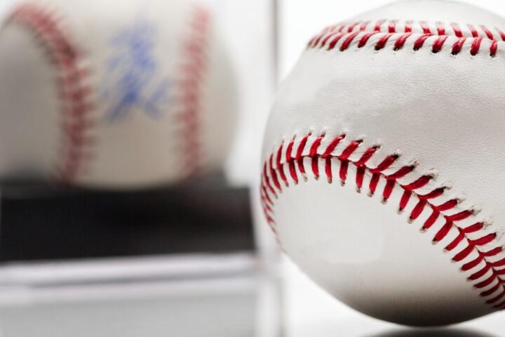 Two baseballs on a reflective white surface; one in focus, the other in a display case blurred in the background.