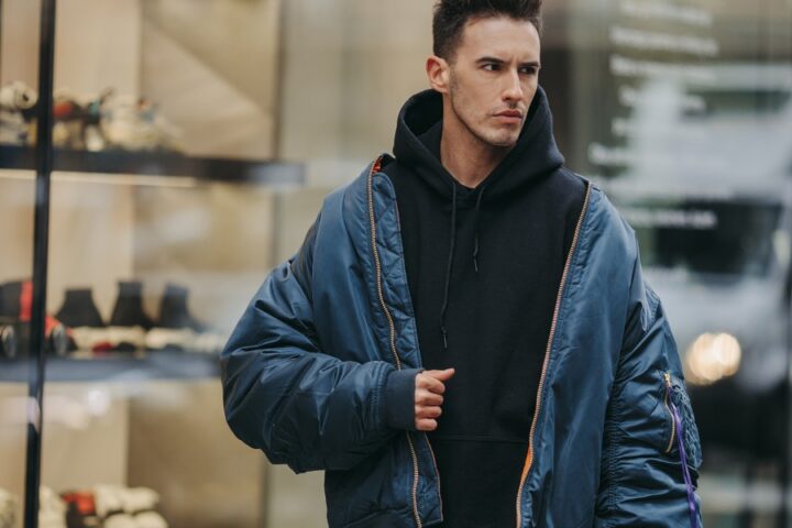 A stern-looking man walking down the street while wearing a black hoodie and a bulky blue jacket with orange zippers.