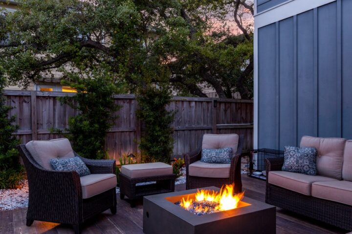 A backyard patio with an outdoor sofa, chairs, and ottomans. There is a fire pit in the seating area and a fence around the yard.