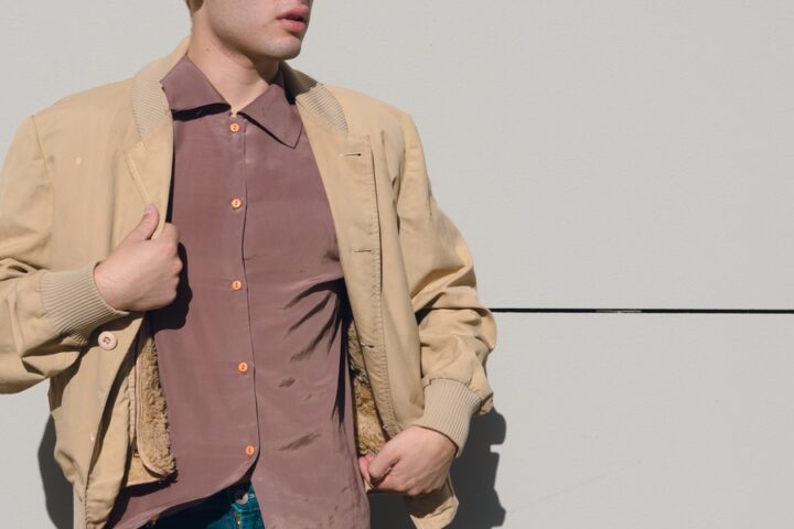 A people wearing blue jeans, a muted red dress shirt, and a tan work jacket with a furred interior lining.