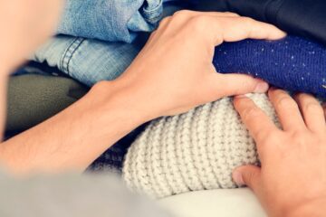 A Caucasian young man sorts through folded clothing on a white shelf, separating a wool shirt from a blue item.