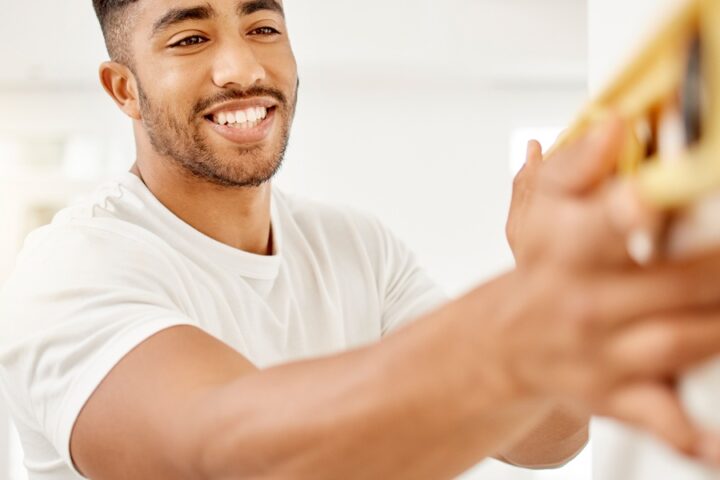 Smiling man holding a yellow spirit level against a wall indoors, checking alignment in a bright room.