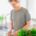 Person preparing a sandwich in a bright kitchen, adding sliced vegetables to the bread with fresh herbs on the counter.